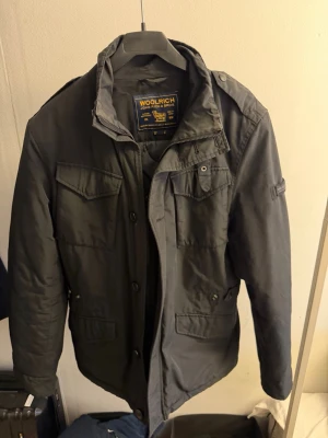 Woolrich jacka  - Snygg svart jacka från Woolrich med flera fickor framtill, axelklaffar och hög krage. Jackan har dragkedja och knappar samt en cool militärinspirerad look. Perfekt för dig som gillar streetwear och funktionella detaljer.