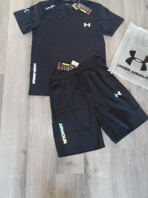 Svart träningsset från Under Armour - Snyggt svart träningsset från Under Armour med t-shirt och shorts. Setet har vita och färgade logotyper på både tröja och byxa. Tillverkat i lätt och snabbtorkande material, perfekt för gym eller löpning. Elastisk midja på shortsen för skön passform. Med regnbågreflektion.