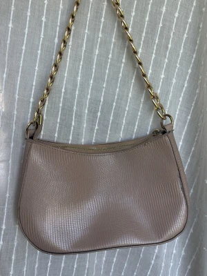 Beige axelremsväska med kedja - Stilren beige handväska i fejkläder med präglat mönster och guldfärgad kedjerem. Väskan har en mjukt rundad form och stängs med dragkedja upptill. Perfekt accessoar för att lyfta din outfit.