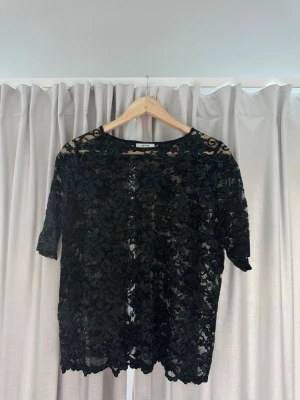 Svart spetsblus med blommönster - Snygg svartgrön från Ganni i genombruten spets med blommönster. Blusen har korta ärmar och rund halsringning. Perfekt att styla med jeans eller kjol för en trendig look.