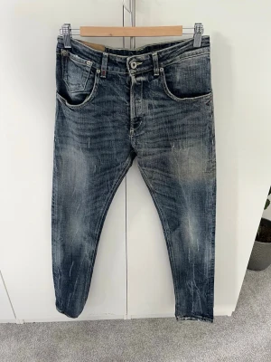Blå slitna jeans från DonDup - Säljer ett par blå jeans från DunDop  med snyggt sliten tvätt och klassisk femficksmodell. Jeansen har orangea sömmar och en diskret logga baktill i midjan. Materialet är denim i bomull och passformen är rak.