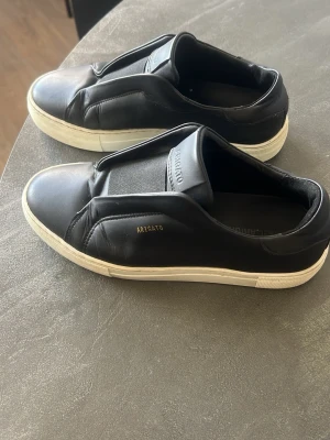 Svarta sneakers från Axel Arigato - Stilrena svarta sneakers från Axel Arigato i mjukt läder med vit sula och guldfärgad logga på sidan. Skorna har en clean slip-on design utan snörning och en låg silhuett som passar till det mesta. Perfekta för dig som gillar minimalistisk och modern stil. Använda fåtal gånger, nytt skick 