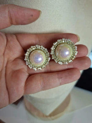Stora pärlörhängen med strass - Säljer ett par statement örhängen med stora vita pärlor i mitten och gnistrande strass runt om. Örhängena har en rund form och guldfärgad metall som bas. Perfekta för dig som vill sticka ut och addera lite extra bling till din look.
