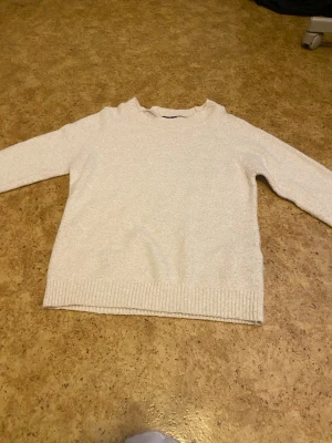 Beige stickad tröja med rund hals - Mysig beige stickad tröja med rund hals och långa ärmar. Tröjan har ribbade muddar vid ärmslut och nederkant, vilket ger en klassisk och enkel look. Perfekt för lager på lager under kyliga dagar. Den är från Vero Moda. Jag vill sälja tröjan eftersom den inte kommer till användning.
