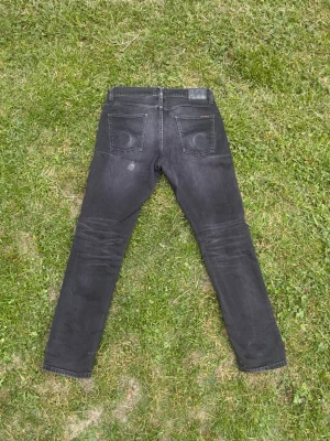 Svarta slim fit jeans från Nudie - Säljer ett par svarta slim fit jeans från Nudie Jeans med klassisk femficksdesign och diskreta slitningar. Jeansen har smal passform och är tillverkade i stretchigt denim för extra komfort. Perfekta för en avslappnad och stilren look. Model Lean Dean.