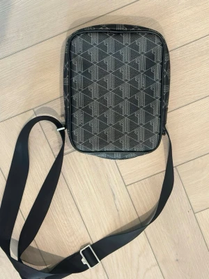 Svart axelväska från Lacoste - Snygg svart axelväska från Lacoste med justerbar rem och ett grafiskt mönster i grått. Väskan har en rektangulär form och ett extra fack framtill med Lacoste-loggan. Perfekt för att bära det viktigaste och ger en stilren look.