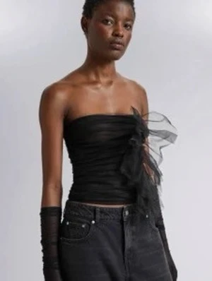 Svart bandeau tubtopp med volang - Trendig svart bandeau tubtopp i transparent mesh med stor volangdetalj framtill. Toppen är axelbandslös och har en tight passform som framhäver siluetten. Perfekt för dig som vill sticka ut med en edgy och modern look.