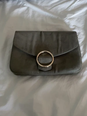 Grå kuvertväska med metallring - Stilren grå kuvertväska i mocka-look med stor rund metallring på locket. Väskan har kedjerem och flera fack inuti för smart förvaring. 