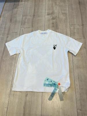 Vit Off-White t-shirt med tryck - Vit t-shirt från Off-White med svart tryck av en hand och texten 'Off' på bröstet. Klassisk rund hals och korta ärmar. Tillverkad i mjuk bomull för en skön och avslappnad look. Perfekt till jeans eller shorts.