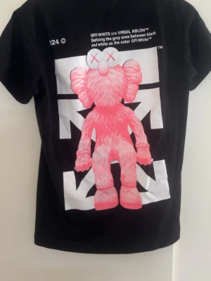 Off-White svart t-shirt med KAWS-print - Svart t-shirt från Off-White med stort rosa KAWS-tryck på ryggen och ett mindre hjärta med Off-White-logga framtill. Klassisk passform och rund hals. Tillverkad i mjuk bomull, perfekt för dig som gillar streetwear och unika prints.