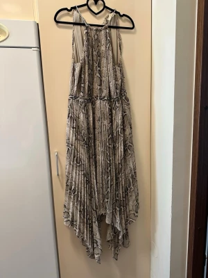 Beige långklänning med ormmönster från Micheal kors - Säljer en beige långklänning med ormmönster och tunna kedjeaxelband. Klänningen har ett luftigt, plisserat tyg och asymmetrisk nederkant som ger en cool och trendig look. Perfekt för dig som vill sticka ut med ett djurmönster och snygga detaljer.