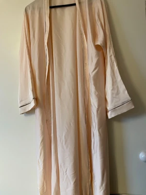 Ljusrosa lång morgonrock / Öppen Abaya - Säljer en elegant ljusrosa morgonrock i ett mjukt, glansigt material. Modellen är lång med vida ärmar och har ett tunt, glittrande kantband vid ärmsluten för en extra lyxig känsla. Perfekt för att chilla hemma i stil. Aldrig använd, passar längder 160-170.  one size fits all. Ända tecknet på användning är att en knapp fattas på toppen, vilket man kan fixa med nål.
