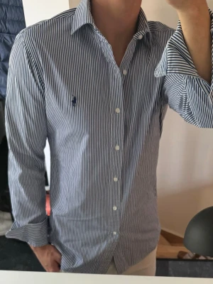 Randig skjorta från Polo Ralph Lauren - Säljer en klassisk blåvit randig skjorta från Polo Ralph Lauren med broderad logga på bröstet. Skjortan har lång ärm, knappar framtill och är tillverkad i bomull. Perfekt för dig som gillar stilrena och tidlösa plagg. Storlek 39 vilket motsvarar m 