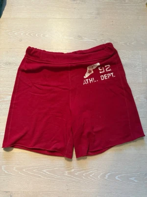 Abercrombie & Fitch Röda sportiga shorts  - Röda shorts med tryck 'ATHL. DEPT. 92' och en vinge framtill. Mjukt material och elastisk midja, perfekt för en avslappnad och sportig look.