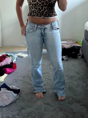 Ljusblå bootcut jeans med rosa detaljer - Säljer ett par ljusblå bootcut jeans med snygga rosa detaljer på hällorna och fickorna. Jeansen har låg midja och klassisk femficksdesign. Perfekta för dig som gillar en avslappnad och trendig stil.