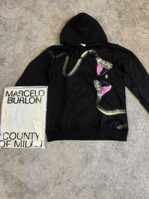 Svart hoodie med ormtryck Marcelo Burlon - Svart hoodie från Marcelo Burlon County of Milan med unikt ormtryck i grönt, silver och rosa på framsidan. Klassisk huva med dragsko och ribbade muddar. Stilen är streetwear och materialet är mjuk bomull. Perfekt för dig som vill sticka ut med en cool design.