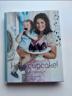Hello cupcake! av Leila Lindholm - En inspirerande och färgglad bok om cupcakes av Leila Lindholm. Omslaget har en bild på Leila och ett barn, där de håller en tallrik med chokladcupcakes. Perfekt för dig som älskar att baka och vill testa nya, kreativa recept på söta bakverk.
