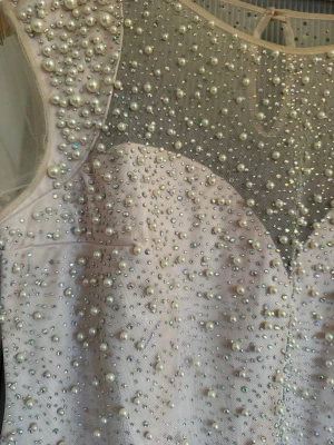 Lång ljusrosa klänning med pärlor - Elegant långklänning i ljusrosa med transparent mesh upptill och ärmar. Klänningen är dekorerad med pärlor och glittriga detaljer över hela framsidan och axlarna. Perfekt för dig som vill sticka ut med en glamorös look.