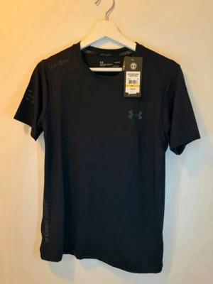 Under armour t shirt  - Snygg svart t-shirt från Under Armour med diskret logga på bröstet och tryck på ärmen. Tillverkad i ett lätt och funktionellt material som passar perfekt för träning. Klassisk rund halsringning och korta ärmar för en sportig look.