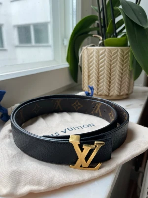 Louis Vuitton monogram bälte svart/guld - Snyggt Louis Vuitton bälte i svart skinn med klassiskt monogrammönster och en stor LV-spänne i guld. Perfekt accessoar för att lyfta din outfit med en lyxig touch. Bältet har en stilren design och är lätt att matcha med olika looks.