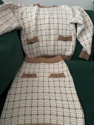 Beige och vit stickad bodyset - Säljer ett snyggt tvådelat set med stickad tröja och kjol i beige och vitt rutmönster. Setet har ribbade muddar, dekorativa fickor med knappar och en mjuk, bekväm känsla. Perfekt för dig som gillar klassisk och trendig stil.