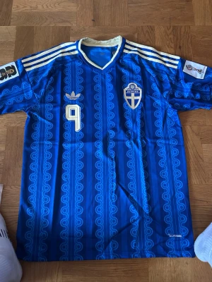 Blå Sverige retro fotbollströja Adidas - Retro blå fotbollströja från Adidas med Sveriges landslagsemblem och nummer 9 på bröstet. Tröjan har vita och gula detaljer, klassiska Adidas-ränder på axlarna och mönstrad struktur i tyget. V-ringad krage i vitt och blått. Perfekt för fotbollsfans!
