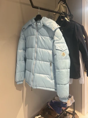 Ljusblå dunjacka från Moncler - Snygg ljusblå dunjacka från Moncler med huva och dragkedja framtill. Jackan har två framfickor med dragkedja, ribbade muddar och Moncler-logga på ärmen. Perfekt för kalla vinterdagar och riktigt trendig färg.