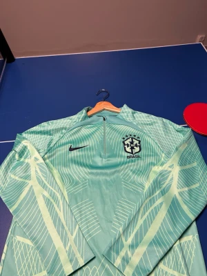 Nike Brasil träningströja mintgrön - Mintgrön långärmad träningströja från Nike med Brasil-logga på bröstet. Tröjan har halv dragkedja och grafiskt mönster i ljusare nyanser. Perfekt för fotbollsträning eller sportiga aktiviteter.