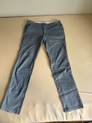 Grå chinos med raka ben från Jack & Jones  - Snygga grå chinos med raka ben och klassisk passform. Byxorna har snedställda fickor fram och två bakfickor med knapp. Perfekta för en stilren och avslappnad look. Materialet känns mjukt och bekvämt.