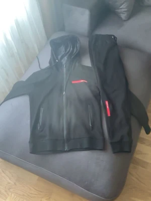 Svart tracksuit-set från Prada - Snyggt svart tracksuit-set från Prada med dragkedja på hoodien och röda detaljer med logga på både jacka och byxa. Setet har huva med snörning och praktiska fickor. Perfekt för en sportig och trendig look. Storleken är M