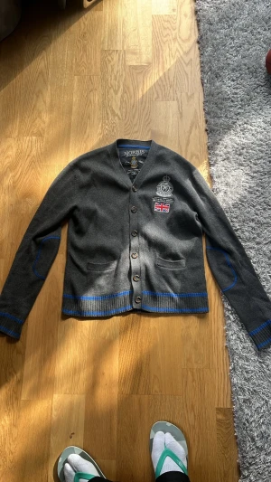 Grå kofta från Morris med brodyr - Snygg grå kofta från Morris med blå detaljer och broderad logga samt brittisk flagga på bröstet. Koftan har knäppning framtill, två fickor och ribbade muddar. Perfekt för en stilren och avslappnad look.