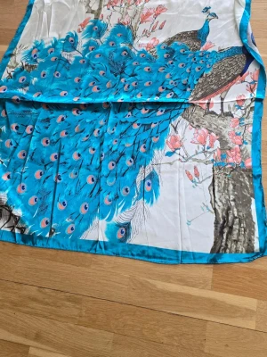 Färgglad sjal med påfågelmotiv - Säljer en stor, fyrkantig sjal i silkesliknande material med ett färgstarkt påfågelmotiv i blått, turkos och inslag av rosa och grönt. Sjalen har tryckt mönster med blommor och naturdetaljer, perfekt för att liva upp din outfit.