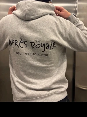 Grå hoodie från Après Royale - Snygg grå hoodie från Après Royale med svart tryck på ryggen där det står 'APRÈS ROYALE' och 'NOLT NORDIC ALPINE'. Hoodien har en klassisk huva och är perfekt för en avslappnad streetstyle-look.