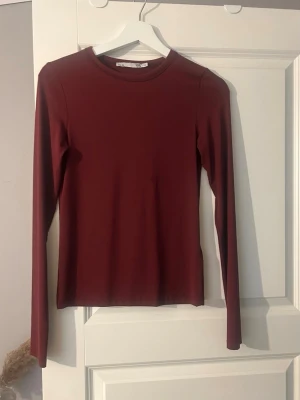 Mörkröd långärmad topp -  Mörkröd långärmad topp med rund halsringning. Toppen har en slim passform och är tillverkad i ett mjukt, stretchigt material som sitter skönt mot kroppen. finns en liten reva på men inget som man tänker på 