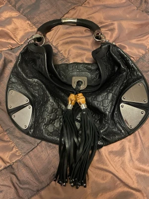 Svart Gucci handväska med tofsar - Cool svart handväska från Gucci i präglat läder med GG-mönster. Väskan har silvriga metalldetaljer, bambuinspirerade detaljer och långa lädertofsar framtill. Handtaget är robust och insidan har ett unikt mönstrat foder samt innerfack med dragkedja.