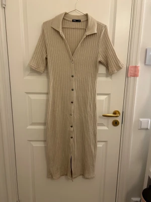 Beige ribbad långklänning från sinsay - Snygg beige långklänning från sinsay med ribbad struktur och knappar hela vägen framtill. Klänningen har korta ärmar, krage och en smickrande v-ringning. Perfekt för dig som gillar en enkel men trendig stil.