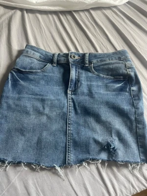 Blå jeanskjol från Pieces - Trendig blå jeanskjol från Pieces i kort modell. Kjolen har rå kant nertill och en liten slitning framtill för en cool look. Klassisk femficksdesign och knappstängning i midjan. Perfekt att matcha med sneakers eller boots.