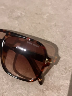 Tom Ford bruna solglasögon - Snygga solglasögon från Tom Ford med brunmelerad båge och stora fyrkantiga glas. Glasen är bruna och bågen har en klassisk aviator-inspirerad form. Perfekt accessoar för att lyfta din stil.