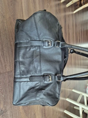 Svart weekendbag i läderlook - Snygg svart weekendbag med två långa handtag och detaljer i läderimitation. Väskan  är slitstark och har dragkedja upptill, innerfack och justerbara spännen på sidorna. Perfekt storlek för kortare resor och enkel att bära över axeln. 
