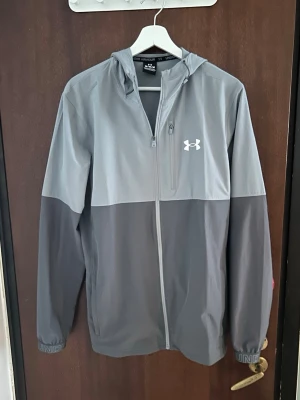 Grå vindjacka från Under Armour - Snygg tvåtonad grå vindjacka från Under Armour med huva och dragkedja framtill. Jackan har en bröstficka med dragkedja och elastiska muddar med logga. Perfekt för blåsiga dagar och sportiga outfits. Pris går att diskutera!!!