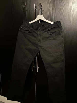 Svarta jeans från Emporio Armani - Säljer ett par svarta jeans från Emporio Armani med klassisk femficksmodell och silverfärgad logga på bakfickan. Jeansen har rak passform och är tillverkade i ett mjukt bomullsmaterial med lite stretch för extra komfort.