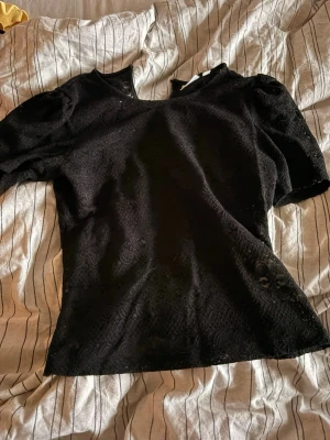 Svart spetsblus med puffärm - Snygg svart blus i spets med puffärmar och rund hals. Blusen har ett genombrutet spetsmönster som ger en elegant look och är lätt figursydd. Perfekt att styla med jeans eller kjol för en trendig outfit.