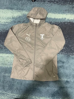 Grå Malmö FF vindjacka från Puma - Snygg grå vindjacka från Puma med Malmö FF-logga och Volkswagen-märke på bröstet. Jackan har huva, hel dragkedja och elastiska muddar vid ärmarna. Perfekt för dig som vill representera ditt lag med stil.