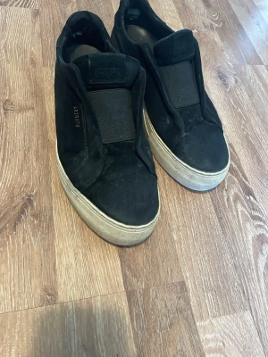 Svarta slip-on sneakers från Axel Arigato - Svarta slip-on sneakers från Axel Arigato med bred resår över vristen och vit, tjock sula. Ovandelen är i mocka och skon har en minimalistisk design utan snörning. Märkeslogga syns på sidan och plösen. Perfekt för en clean och stilren look. Liten skada på hälen men annars inga andra defekter.