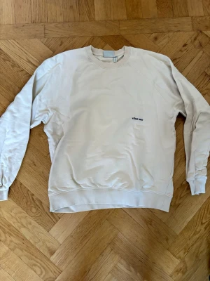Vit sweatshirt från Yoke - Vit sweatshirt från Yoke med rund halsringning och diskret svart broderad logga på bröstet. Tillverkad i mjuk bomull och har ribbade muddar vid ärmslut och nederkant. Enkel och clean design som passar till det mesta.