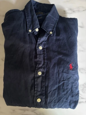 Marinblå skjorta från Polo Ralph Lauren - Klassisk marinblå skjorta från Polo Ralph Lauren med knappar framtill och ikonisk röd broderad logga på bröstet. Skjortan har button-down krage och är tillverkad i mjukt bomullstyg. Perfekt för en stilren och avslappnad look.