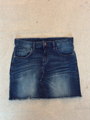 Mörkblå jeanskjol med rå kant - Trendig mörkblå jeanskjol i minilängd med slitna detaljer och rå, fransig kant nedtill. Kjolen har klassiska fem fickor, knapp och dragkedja framtill samt bälteshällor. Perfekt för en avslappnad och cool stil. 