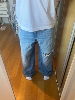 Blåa baggy jeans med hål - Säljer ett par ljusblå baggy jeans med slitningar och ett stort hål på ena knät. Jeansen har raka, vida ben och klassiska fem fickor. Perfekta för en avslappnad och trendig look. Materialet är denim i bomull.