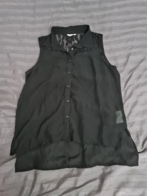 Svart ärmlös blus med spetsdetaljer - Snygg svart ärmlös blus från Cubus med transparent spets upptill och vid axlarna. Blusen har en lätt v-ringning och är gjord i ett tunt, luftigt material som ger en elegant look. Perfekt att styla med jeans eller kjol för en trendig outfit.