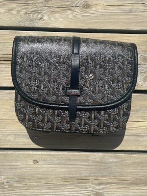 Svart handväska med mönster - Snygg svart handväska med det klassiska vita och gråa mönstret. Väskan har rundade kanter, lock med magnetlås och en svart läderrem som går över locket. Perfekt för dig som gillar exklusiva accessoarer och tidlös design.
